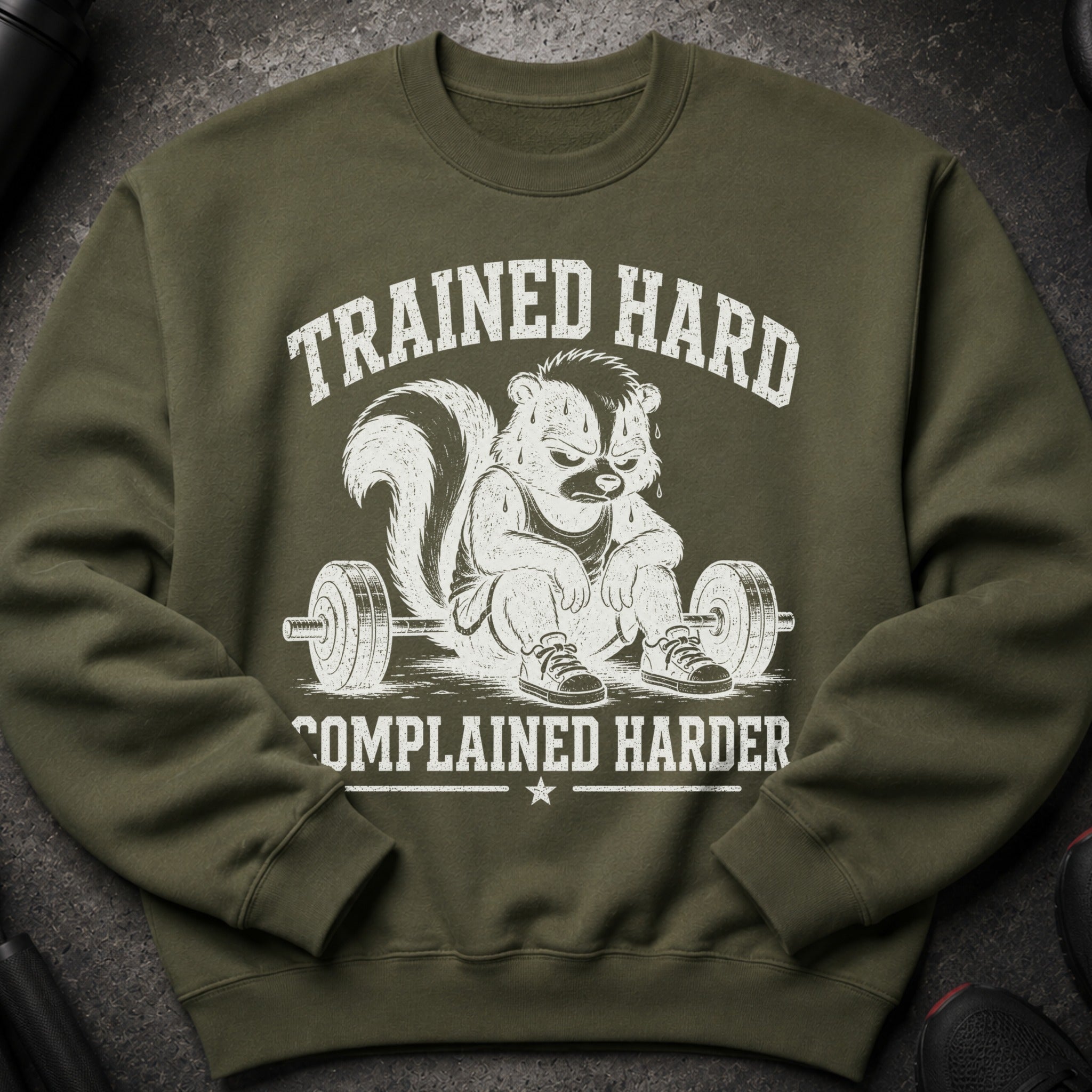 Olive green sweatshirt with distressed graphic of skunk sitting on barbell, text: "TRAINED HARD COMPLAINED HARDER".