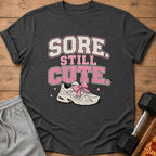 Dark gray t-shirt with text "SORE STILL CUTE" and a sneaker with a pink bow.