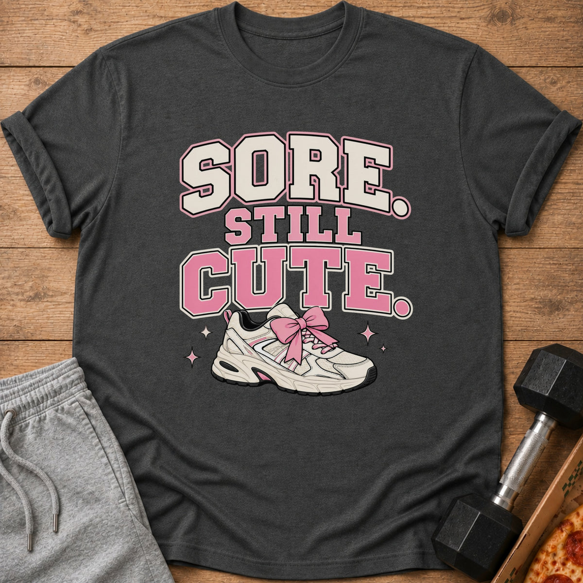 Dark gray t-shirt with text "SORE STILL CUTE" and a sneaker with a pink bow.