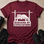 Maroon t-shirt with graphic of a cat on a barbell, reading "Benching My Expectations."