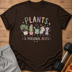 Brown t-shirt with cartoon plants, a watering can, a sneaker, and a dumbbell with text.