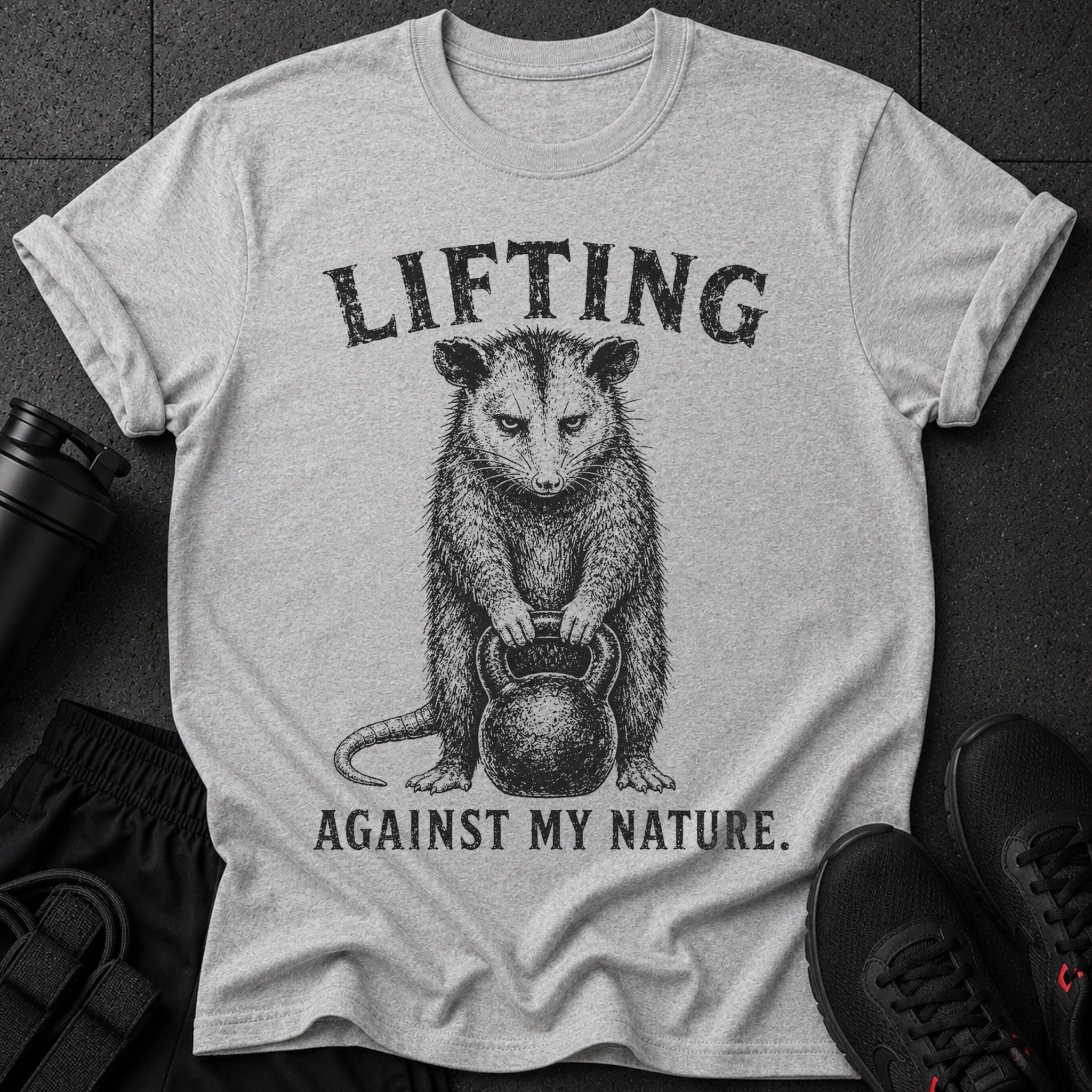 Grey t-shirt with possum holding kettlebell and text "LIFTING AGAINST MY NATURE."