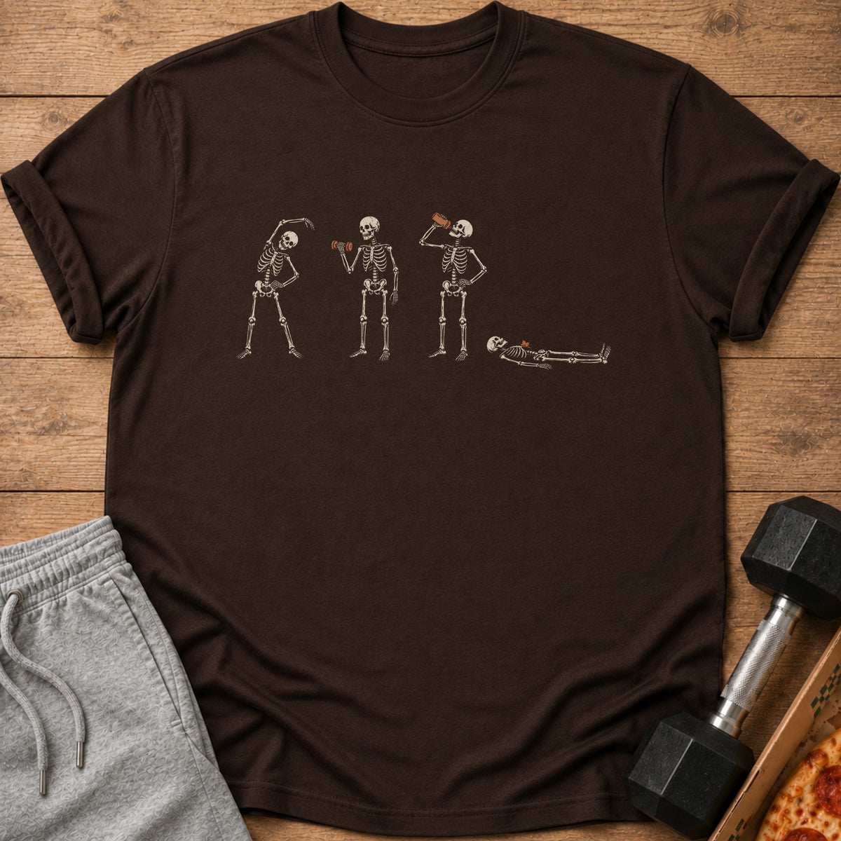 Dark brown t-shirt with four skeletons: stretching, lifting weights, drinking, and resting.