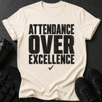 Light beige t-shirt with black text reading "ATTENDANCE OVER EXCELLENCE" and a checkmark.