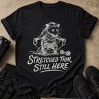 Black t-shirt with a distressed print of a possum working out with resistance bands and dumbbells, text says "Stretched Thin, Still Here".