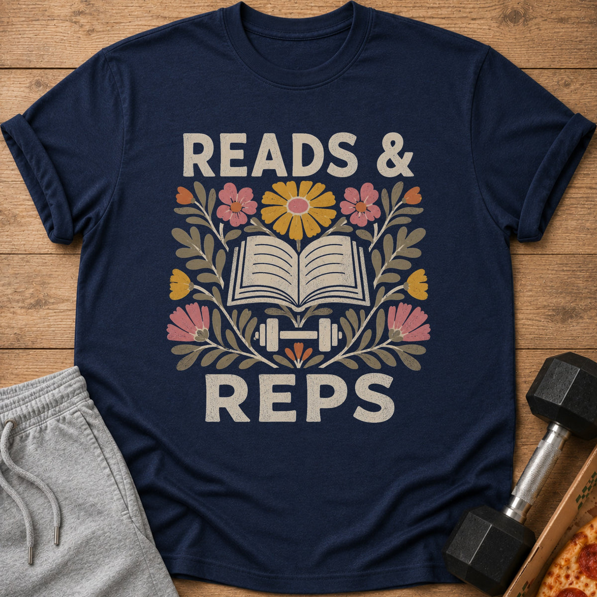 Navy blue t-shirt featuring floral illustrations, an open book, a dumbbell, and the text READS & REPS.