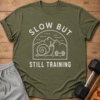 Olive green t-shirt with "SLOW BUT STILL TRAINING" text and snail lifting dumbbell graphic.
