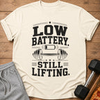 Cream t-shirt with "LOW BATTERY. STILL LIFTING." text, barbell, and battery icon.
