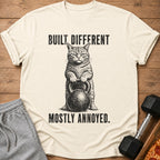Shirt with black graphic of a cat holding a kettlebell and text: "BUILT DIFFERENT. MOSTLY ANNOYED."