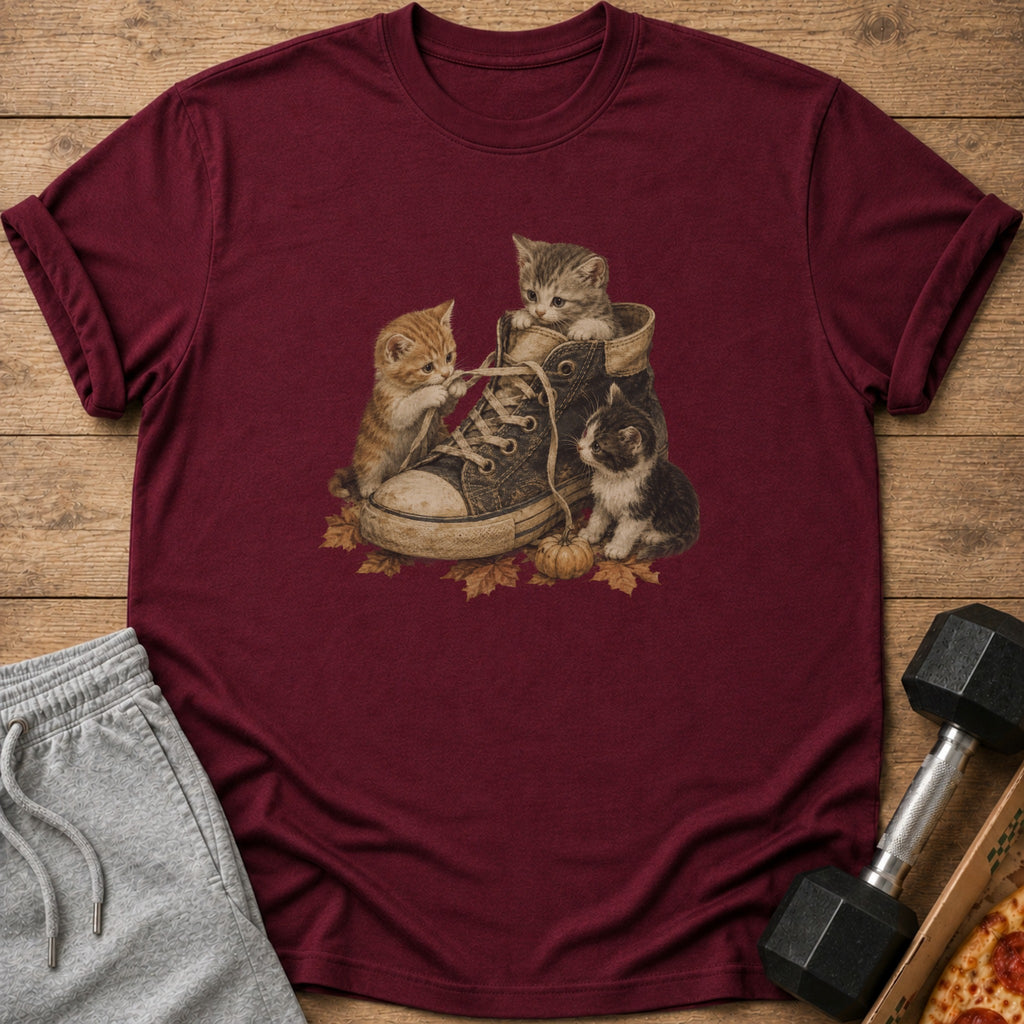 Three kittens play with a shoe on a maroon t-shirt with autumn leaves and a pumpkin.