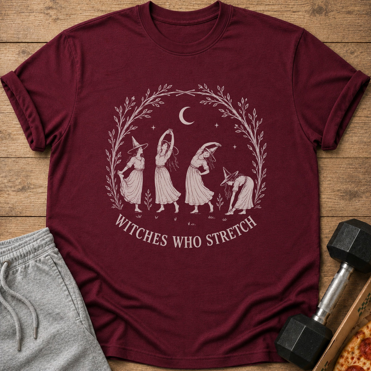 Maroon t-shirt with a graphic of four witches stretching inside a laurel wreath.
