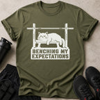 Olive green t-shirt with white graphic of cat bench pressing and text "BENCHING MY EXPECTATIONS".