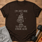 Brown t-shirt with white text and skeleton graphic that says "I'M JUST HERE TO KEEP THE STREAK ALIVE".