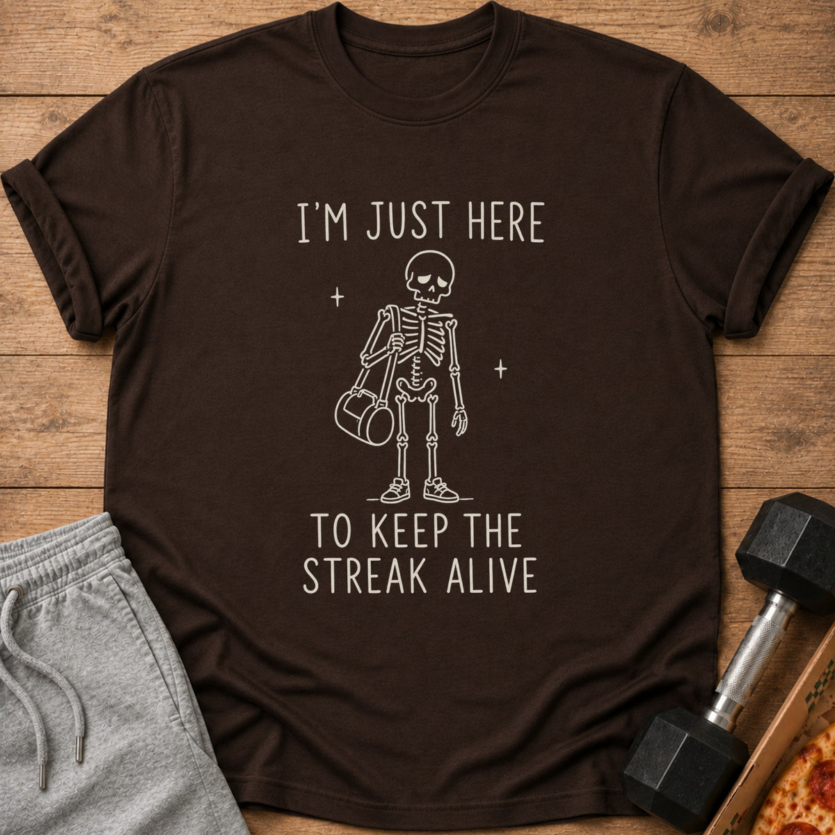 Brown t-shirt with white text and skeleton graphic that says "I'M JUST HERE TO KEEP THE STREAK ALIVE".