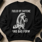 Black sweatshirt with a raccoon lifting weights and holding coffee, "Fueled by Caffeine and Bad Form" text.