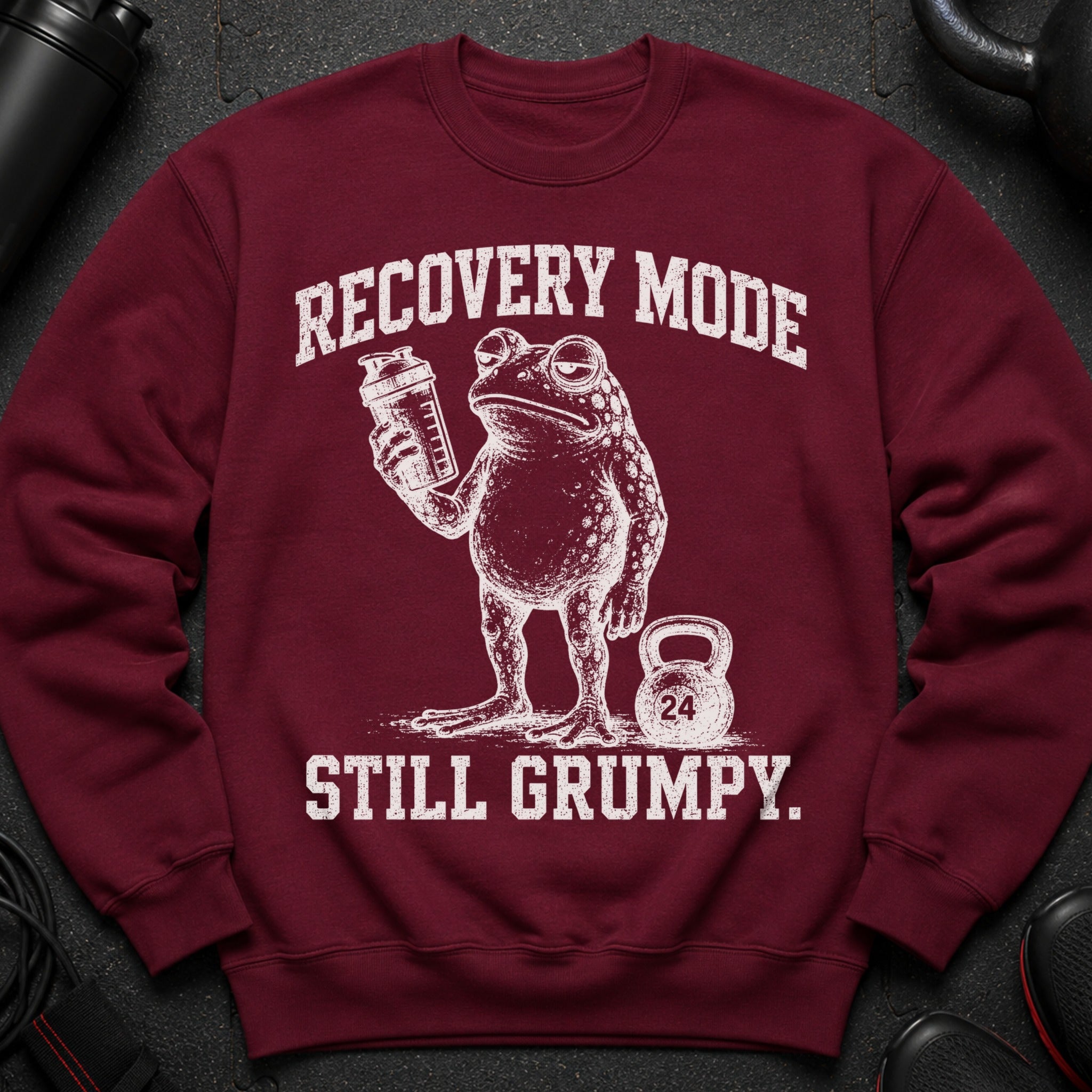 Maroon sweatshirt with distressed print of a grumpy frog holding a shaker and a kettlebell labeled 24.