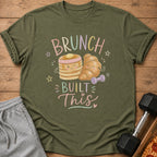 Olive green t-shirt with a graphic of cute cartoon pancakes and a croissant, text reading Brunch Built This, gray sweatpants, and a dumbbell on a wooden background.