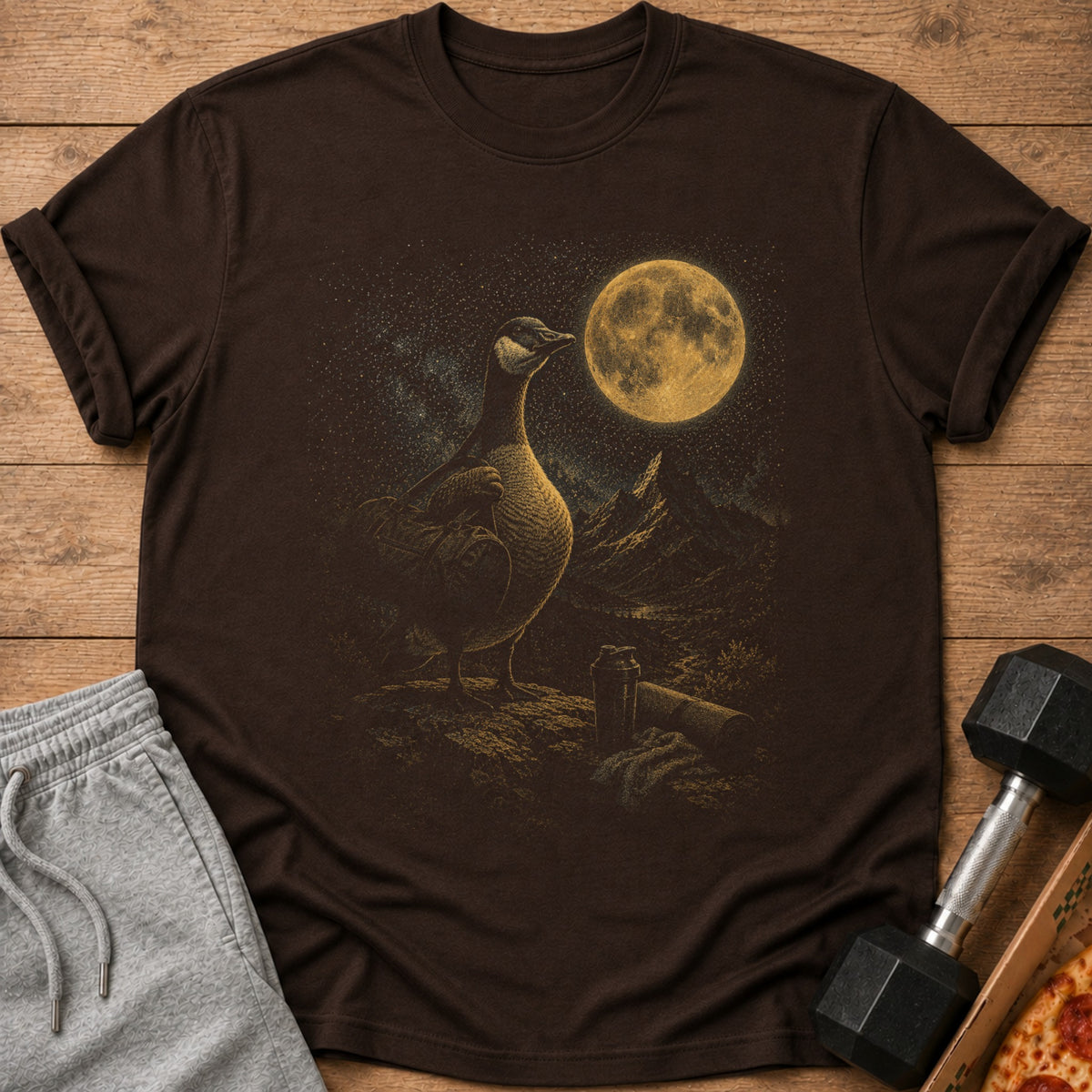Brown t-shirt with a goose and moon graphic, next to grey shorts and a dumbbell.