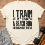 Cream t-shirt with black text reading "I TRAIN LIKE I HAVE A BEACH BODY HIDING SOMEWHERE".