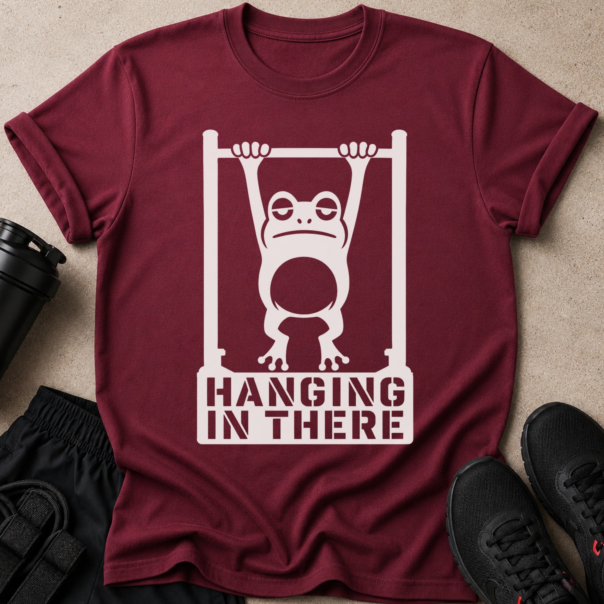 Maroon t-shirt with distressed white graphic of frog hanging from bar and text "HANGING IN THERE".