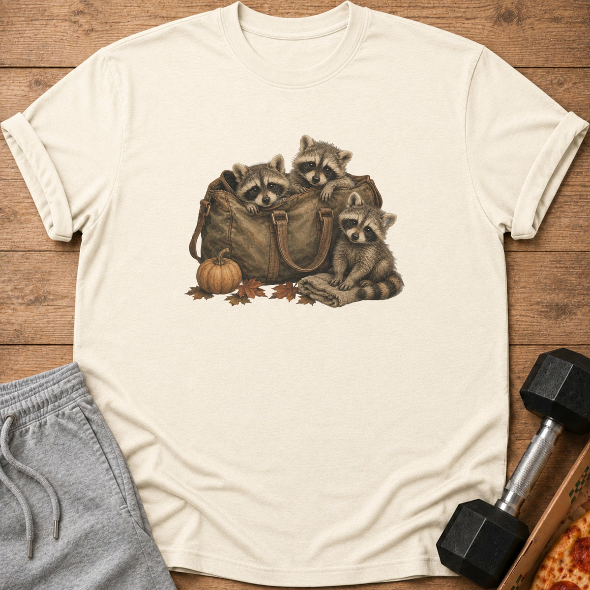 Three raccoons peek out of a duffel bag, with a pumpkin and leaves nearby.