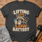 T-shirt with raccoon holding dumbbell and protein shaker, text "LIFTING WITH LOW BATTERY".