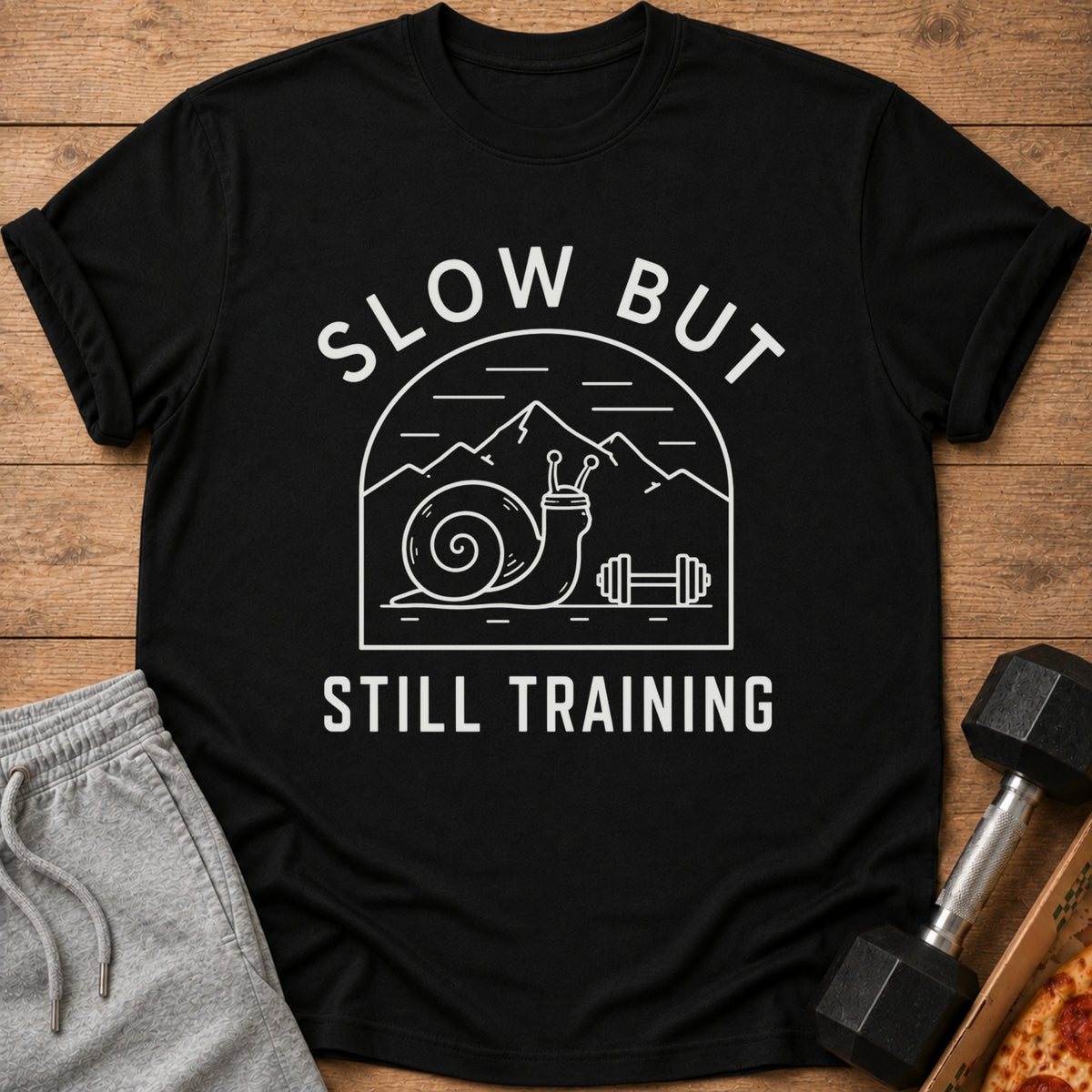 Black t-shirt with white text and graphics of a snail and mountains, reading "SLOW BUT STILL TRAINING".
