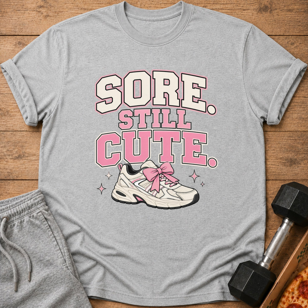 Grey t-shirt with "Sore Still Cute" text and a sneaker with pink bow.