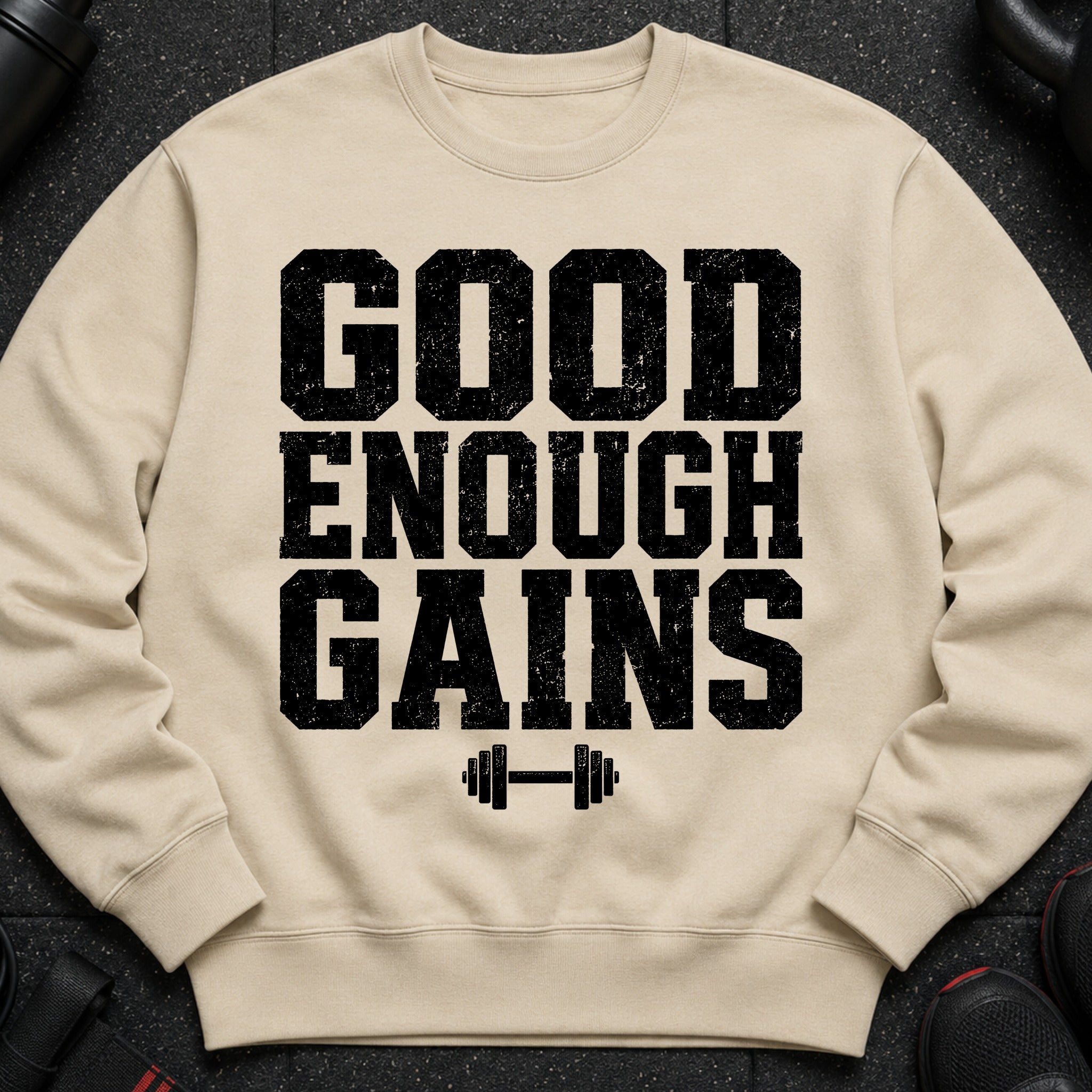 Light tan crewneck sweatshirt with black text "GOOD ENOUGH GAINS" and dumbbell graphic.