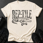 Cream t-shirt with black "REP-TILE" text and alligator lifting dumbbell graphic.