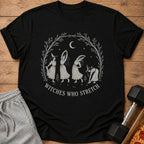 Black t-shirt with a circular design of witches stretching under a crescent moon and stars.
