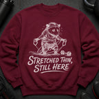 Maroon sweatshirt with a white distressed graphic of a possum exercising and the text "Stretched Thin, Still Here."