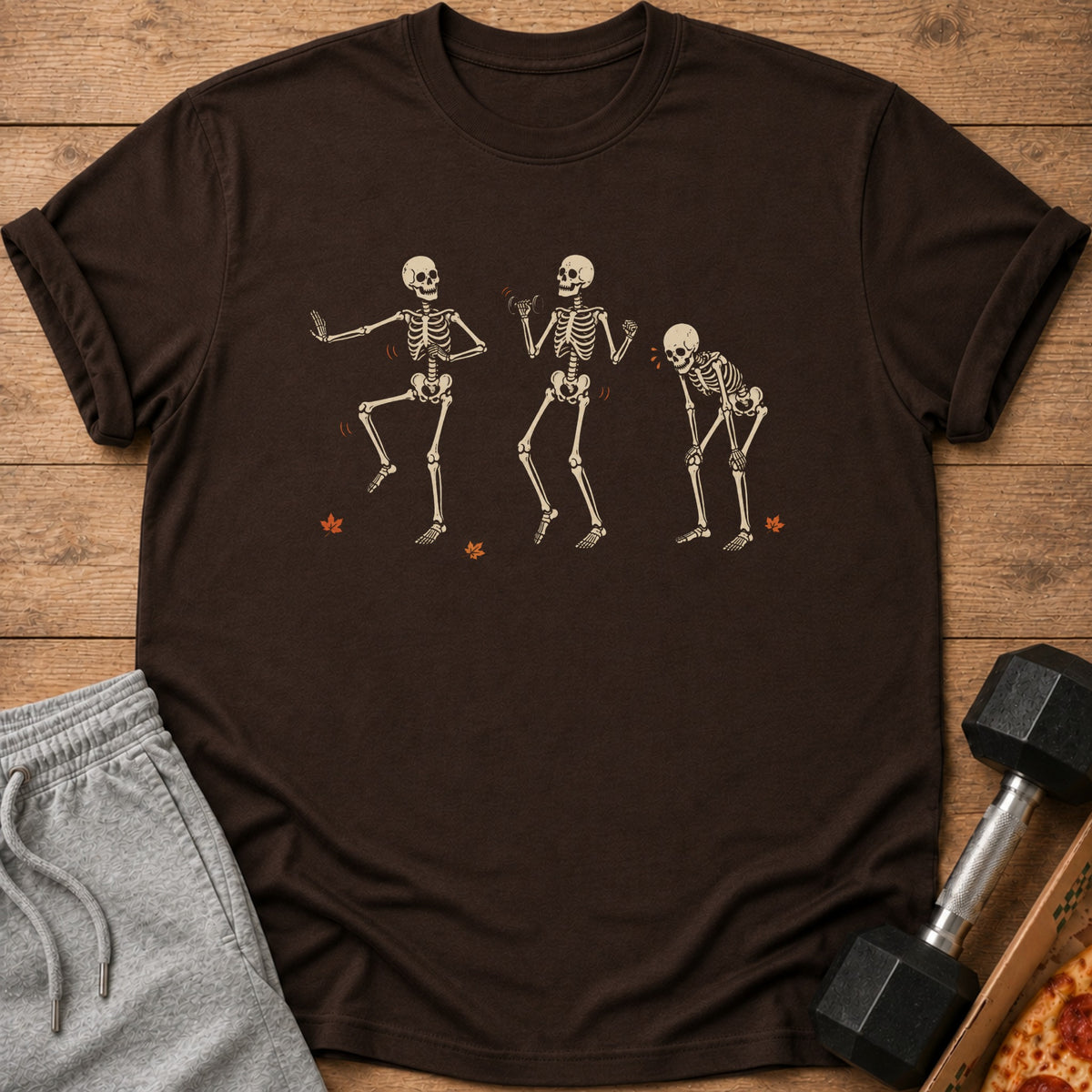 Brown t-shirt with three dancing skeletons and fallen leaves graphic.