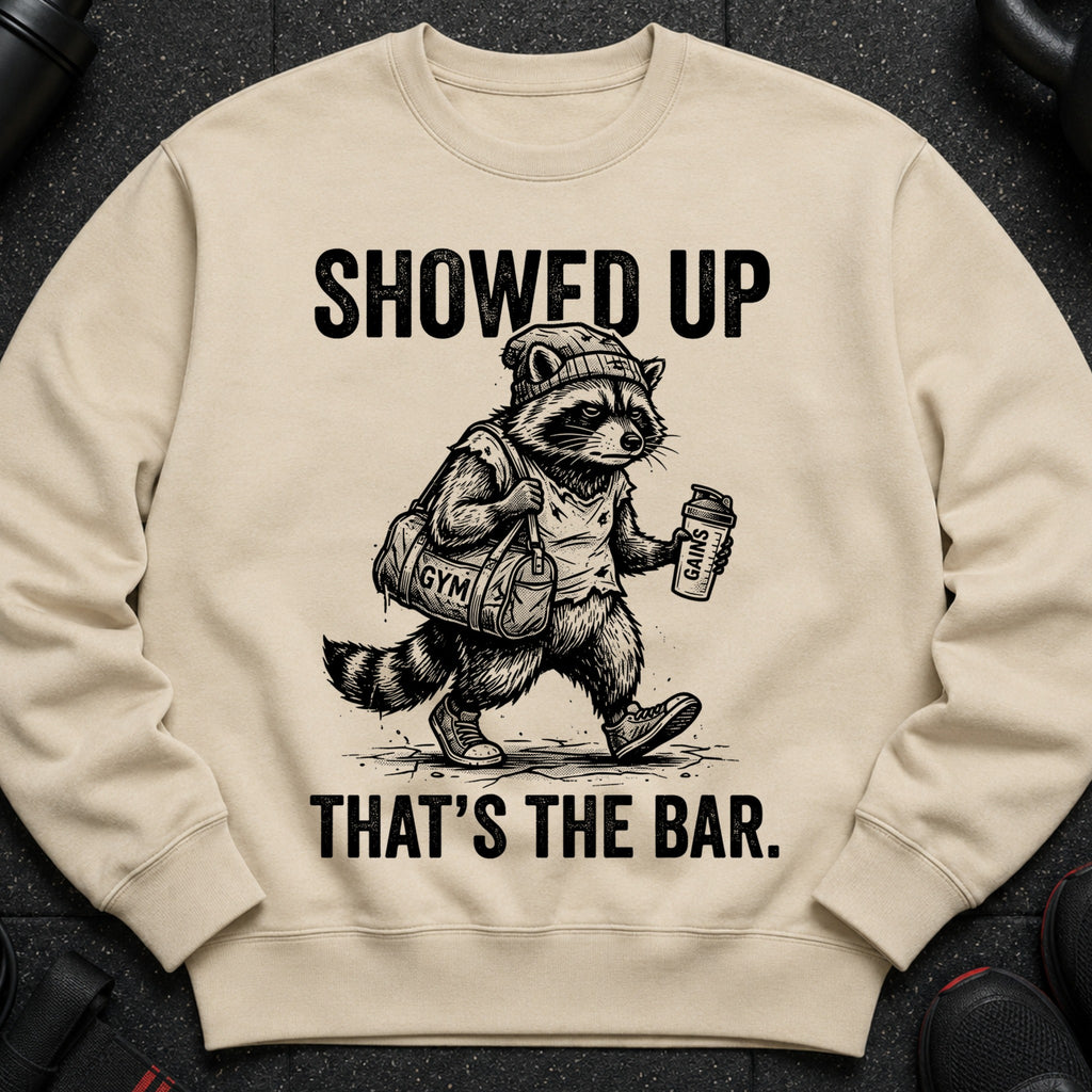 T-shirt with a raccoon mascot holding a gym bag and shaker, with text "SHOWED UP THAT'S THE BAR."