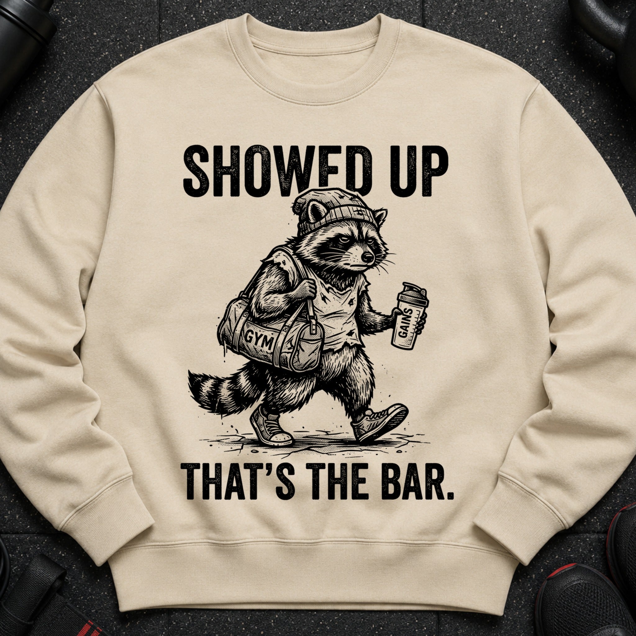 T-shirt with a raccoon mascot holding a gym bag and shaker, with text "SHOWED UP THAT'S THE BAR."