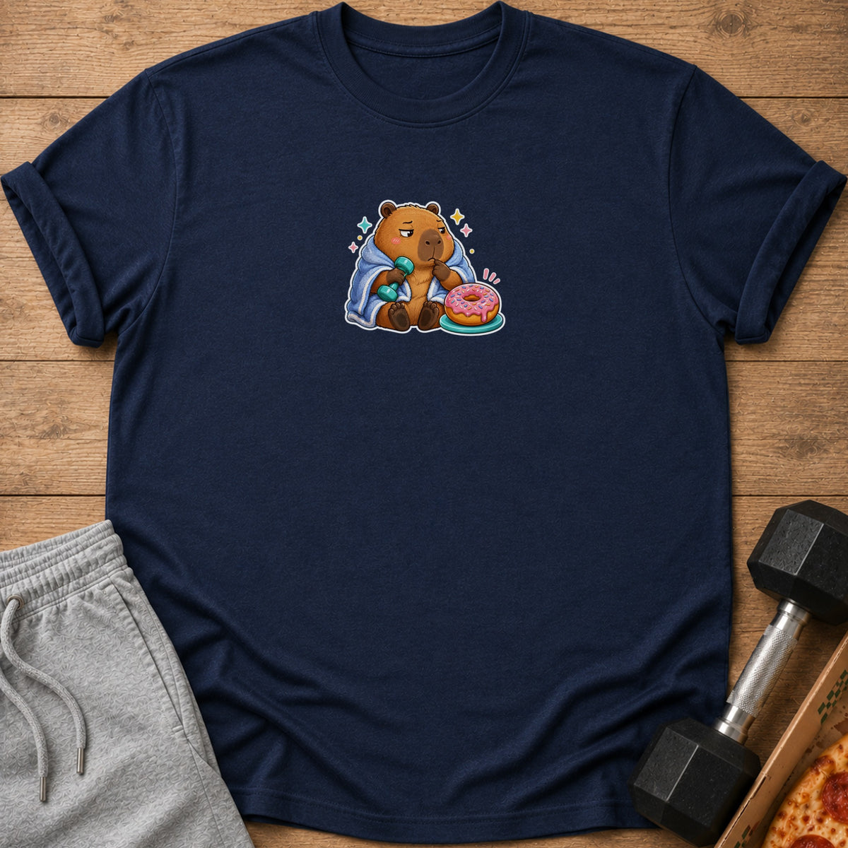 Navy t-shirt with a cartoon capybara wrapped in a blanket holding a dumbbell and a donut.