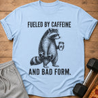 Light blue t-shirt with a raccoon lifting weights and holding coffee, "Fueled by Caffeine And Bad Form."