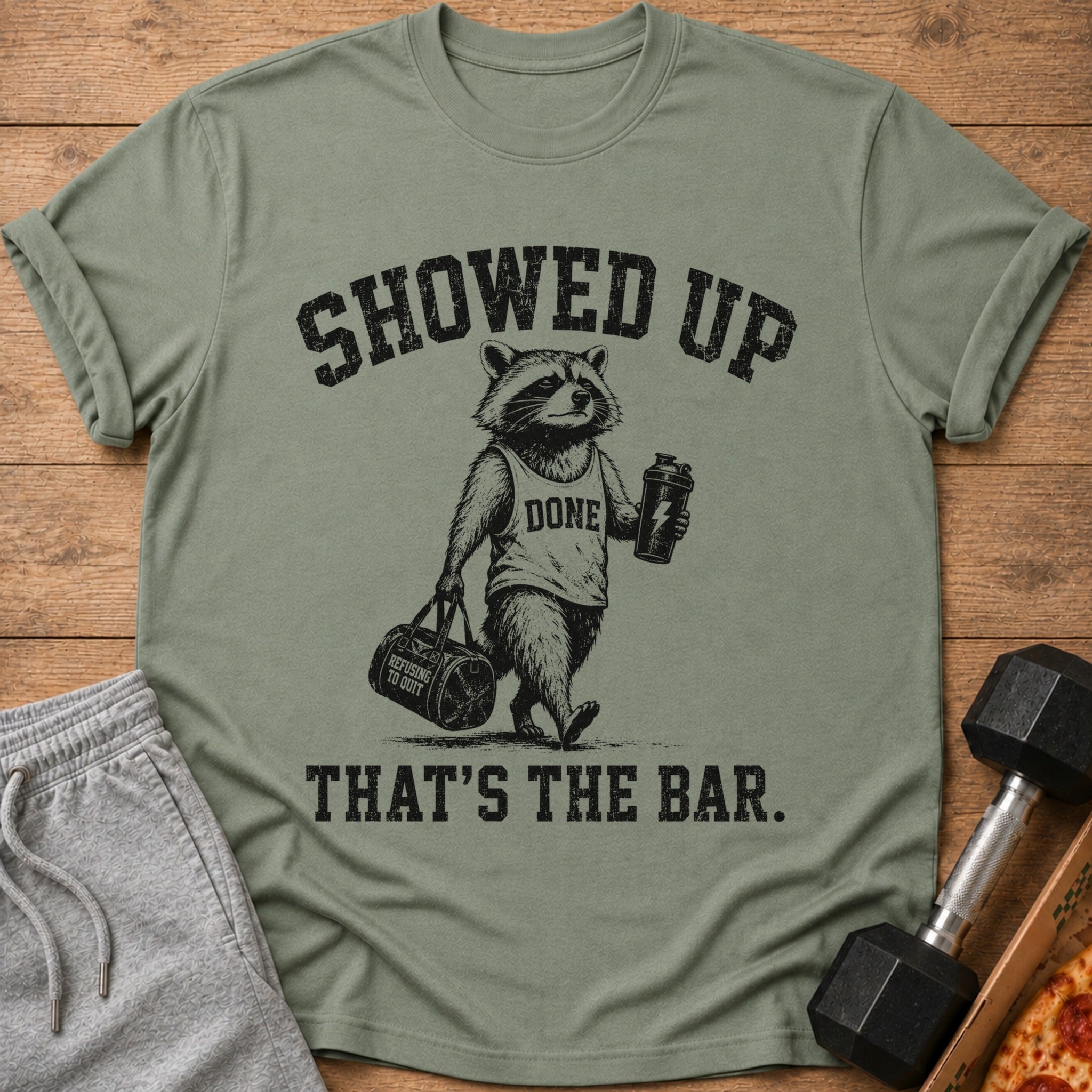Green t-shirt with raccoon graphic and text "SHOWED UP DONE REFUSING TO QUIT THAT'S THE BAR."