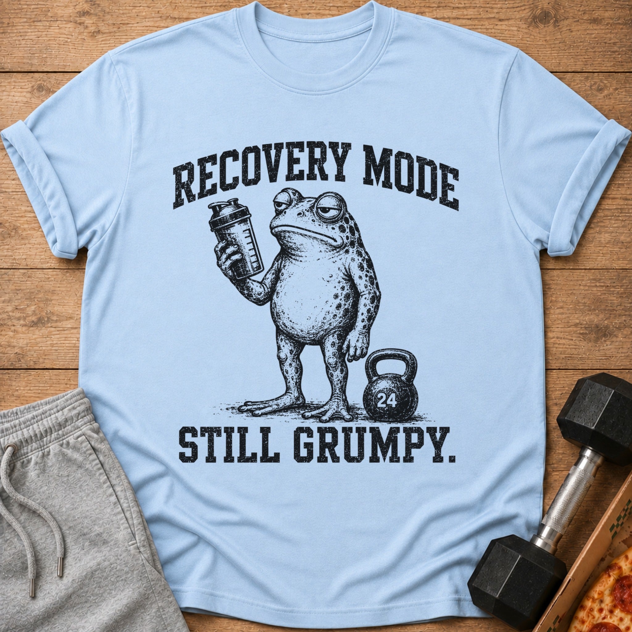 Light blue t-shirt with a grumpy frog holding a shaker bottle and text "RECOVERY MODE STILL GRUMPY."