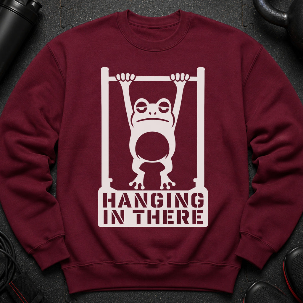 Burgundy sweatshirt with white graphic of frog hanging on bar, text "HANGING IN THERE".