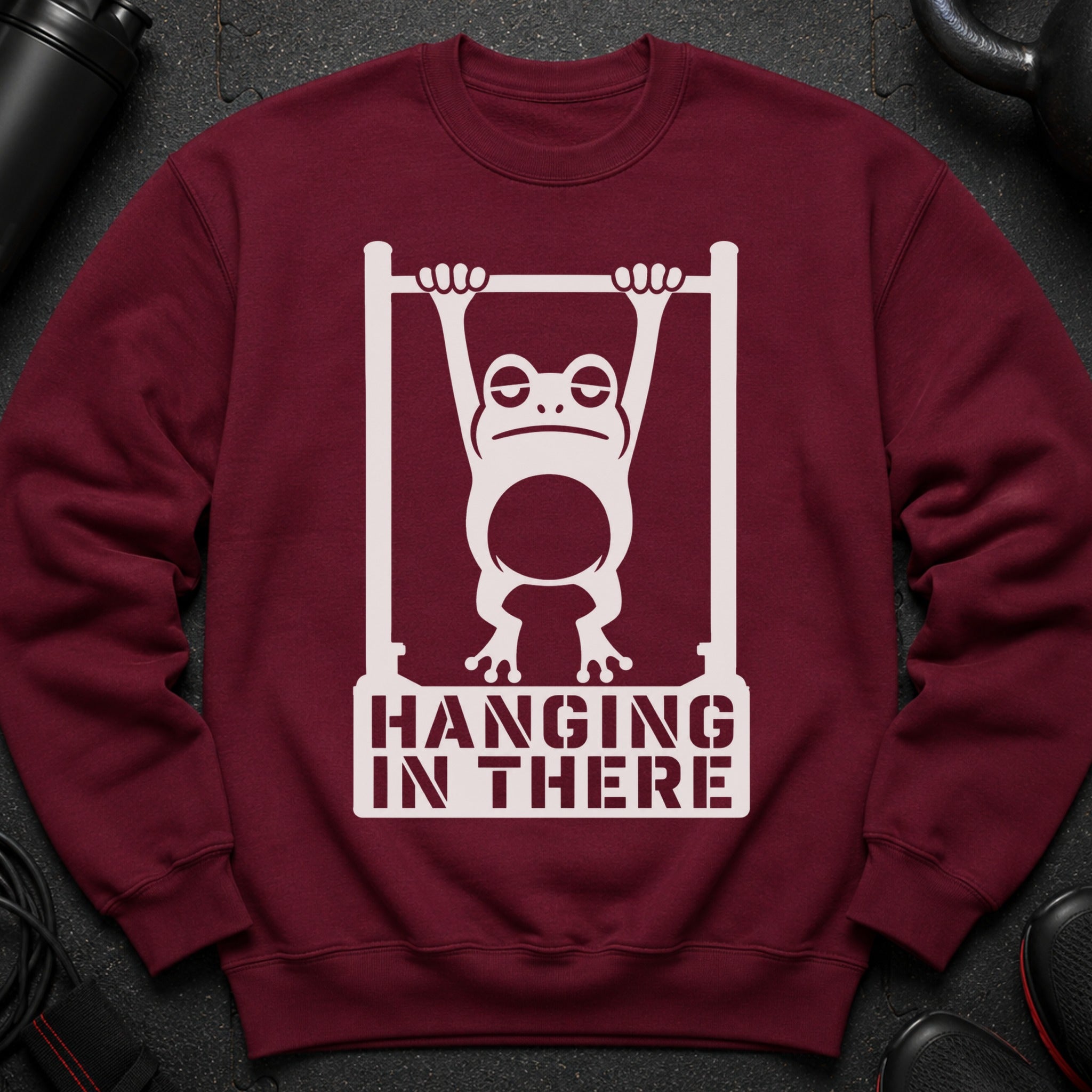 Burgundy sweatshirt with white graphic of frog hanging on bar, text "HANGING IN THERE".