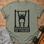 T-shirt with black graphic of frog hanging on bar with text "HANGING IN THERE".
