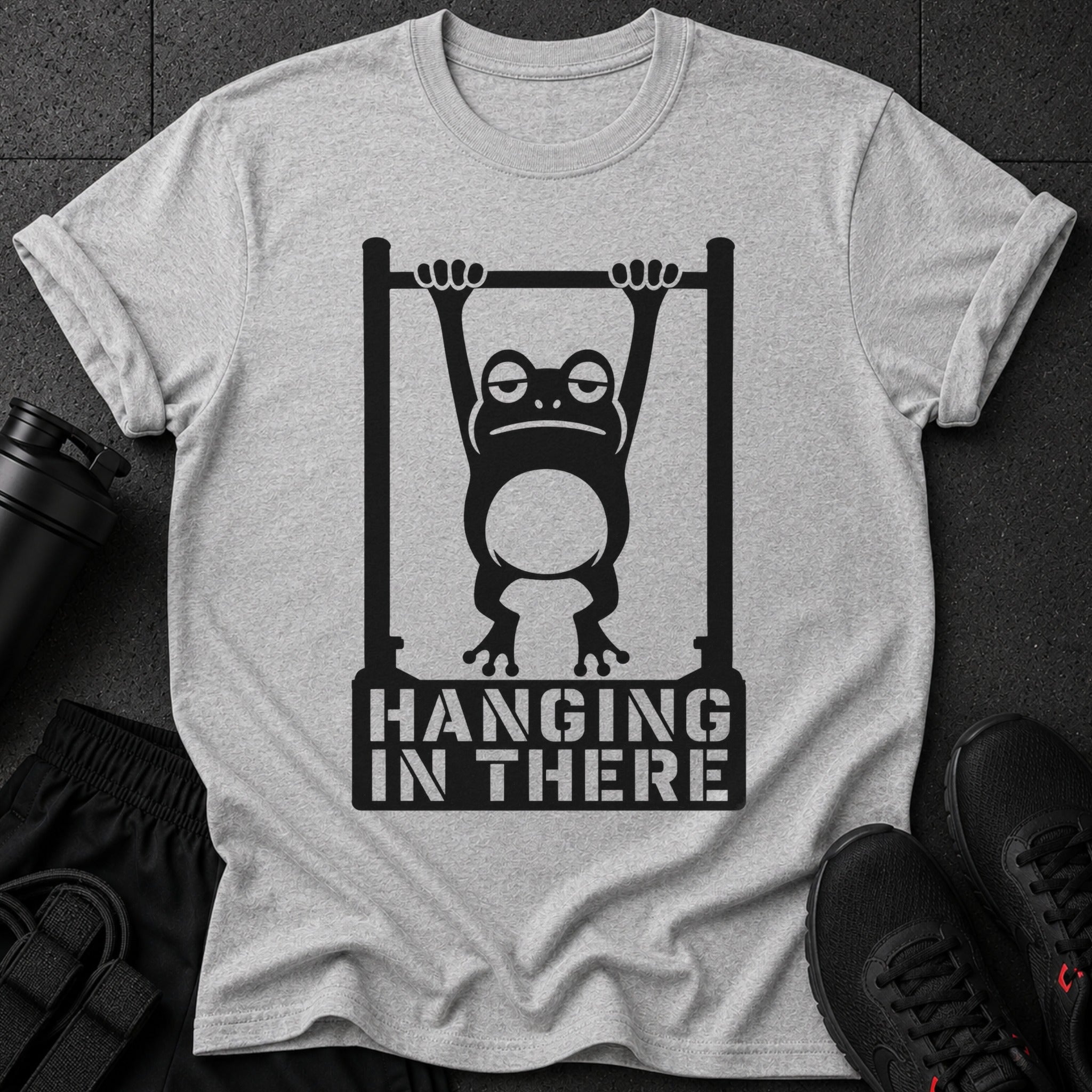 Graphic of a frog hanging from a bar with text "HANGING IN THERE" on a grey t-shirt.