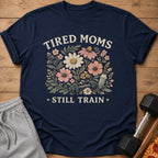 Navy t-shirt with floral design, dumbbell, shaker bottle, and text "TIRED MOMS STILL TRAIN".