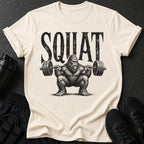 T-shirt with black distressed text "SQUAT" above a hairy Bigfoot lifting a barbell.