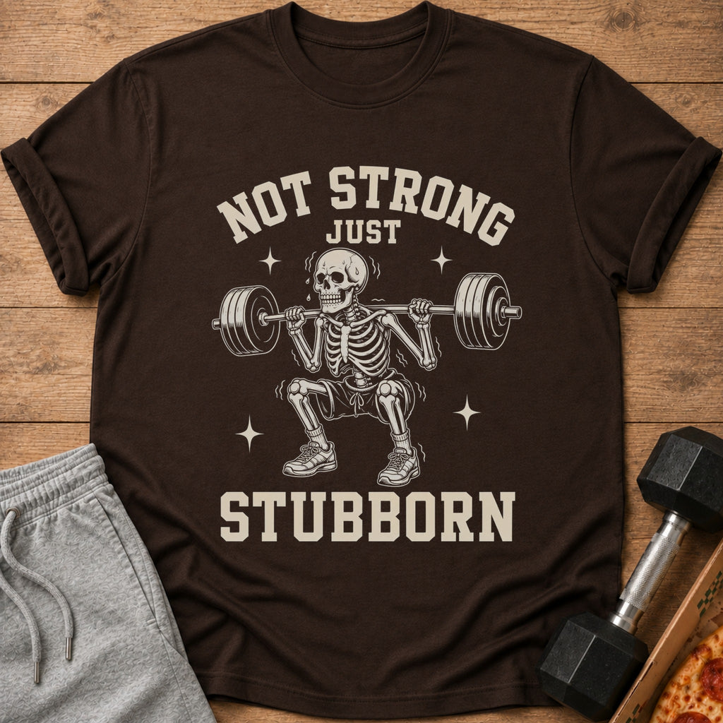 Brown t-shirt with a skeleton lifting weights and the text "Not Strong Just Stubborn".