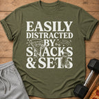 Olive green t-shirt with "Easily Distracted By Snacks & Sets" graphic.