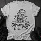 Grey t-shirt with graphic of a possum exercising with resistance bands and text "STRETCHED THIN, STILL HERE".