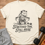 T-shirt graphic of a sweating opossum tangled in resistance bands, with text "Stretched Thin, Still Here".