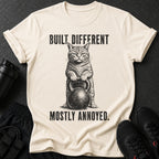 Cream t-shirt with black graphic of a cat holding a kettlebell and text "BUILT DIFFERENT MOSTLY ANNOYED."